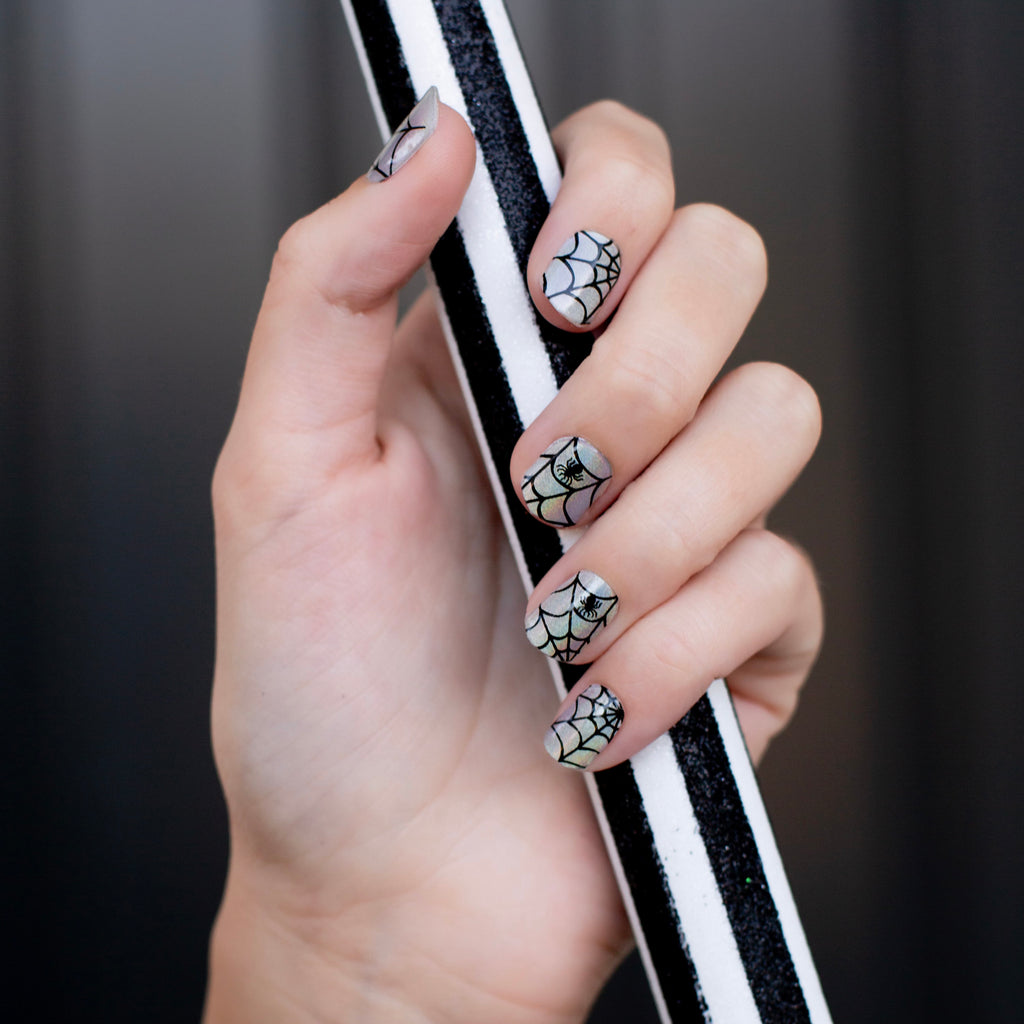 Model's hand flaunting Arachnid Elegance gel nail strips, showcasing the silver-blue iridescent base and intricate black spiderweb designs for perfect spiderweb nails.