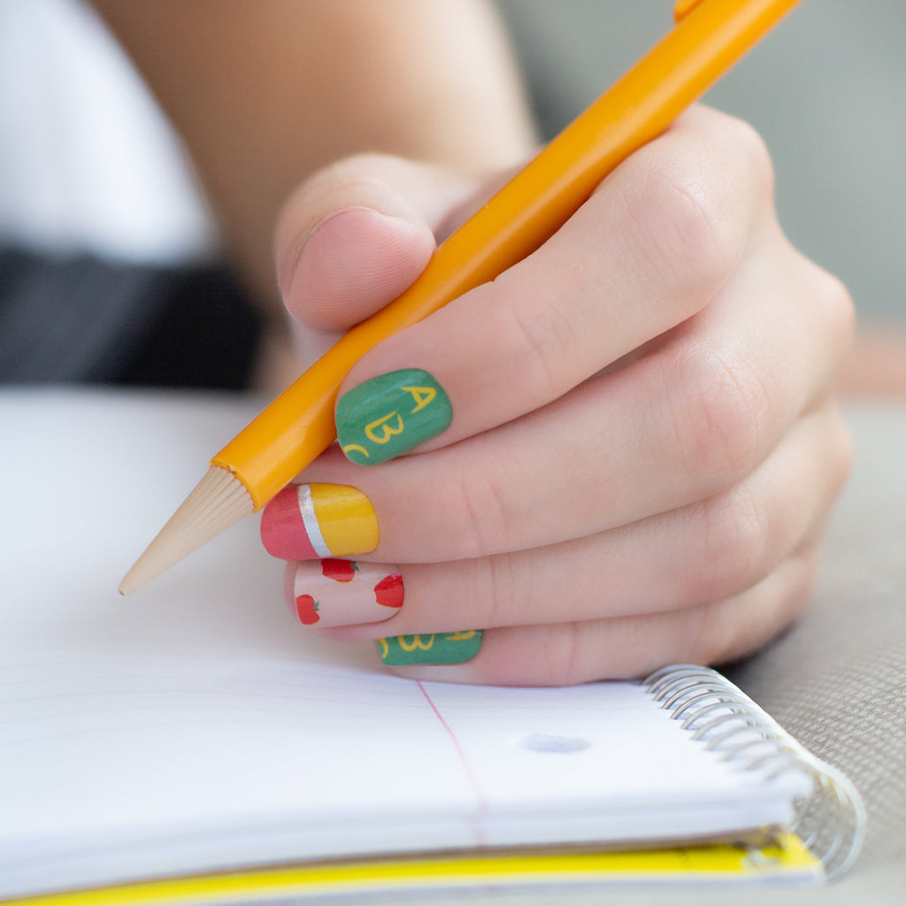 Photo of a model's hand holding a pen and writing on lined paper while featuring 'Apple for the Teacher' nail wraps. The nail wraps have a tan base with lined paper graphics, red apples, yellow pencils, and the ABCs. The design is visible on the nails, and the hand is positioned to show off the nails while writing on the paper.