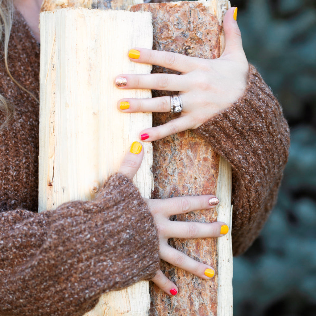 A model's hand holding a bundle of wood, with "Harvest Glitter" nail wraps featured in the foreground, showcasing a mixed manicure with yellow and red nails, along with a stunning gold glitter accent nail, adding a touch of glamour to the overall look.