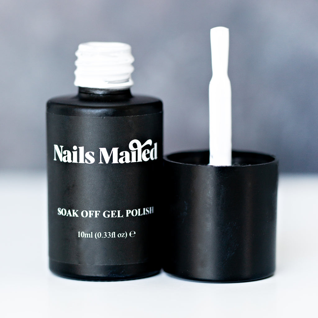 A round bottle of White gel nail polish is shown against a white background. The black label features the words 'Nails Mailed' in white font. The glossy white polish is visible through the clear glass bottle, which has a black cap with a brush applicator sticking out of it.