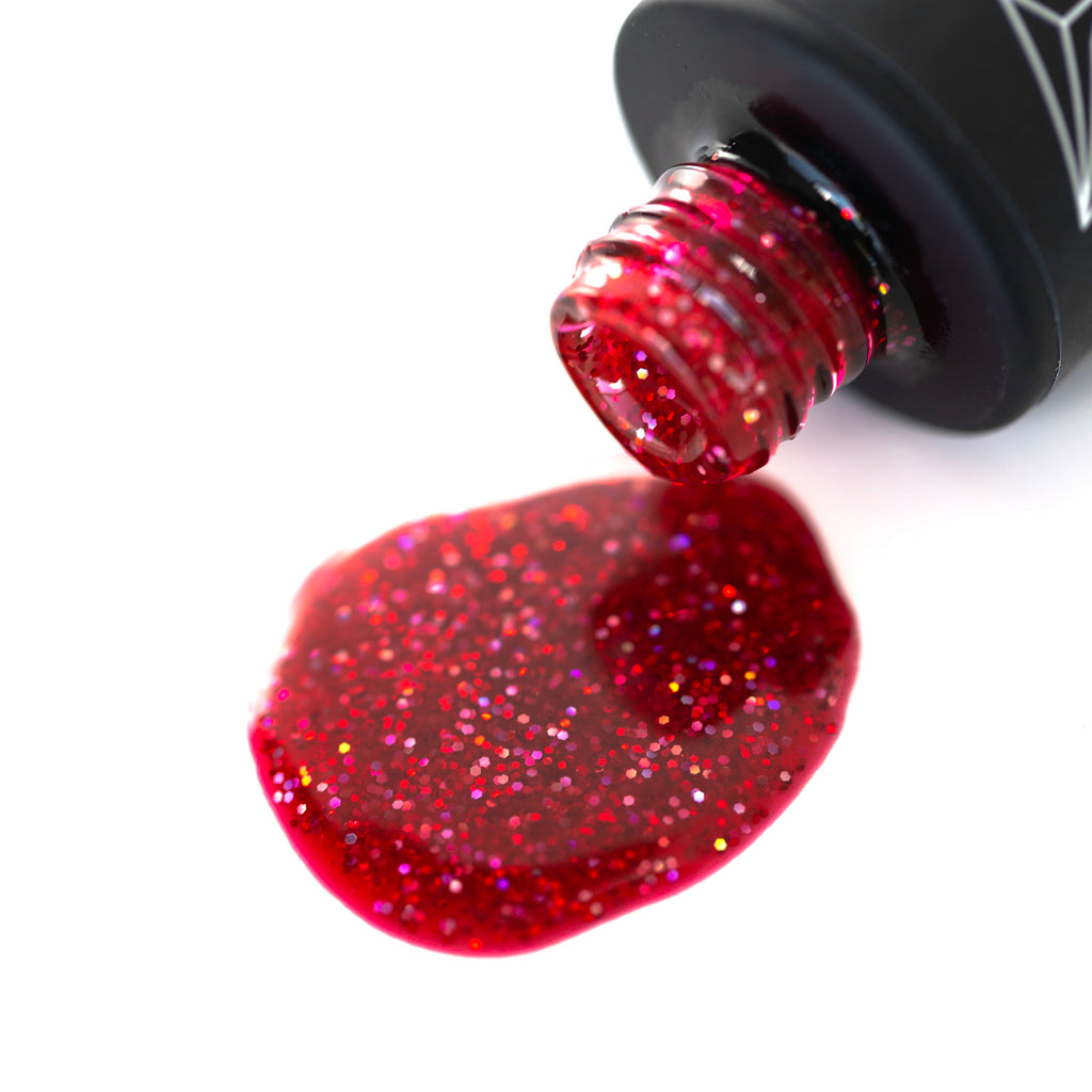 Prom Night gel nail polish, a bright red shade with colorful glitter, is pictured against a white background. The round bottle has a black cap with a brush applicator sticking out of it. The light reflects off the glittery surface of the polish, highlighting the fiery red base with playful colorful accents. 